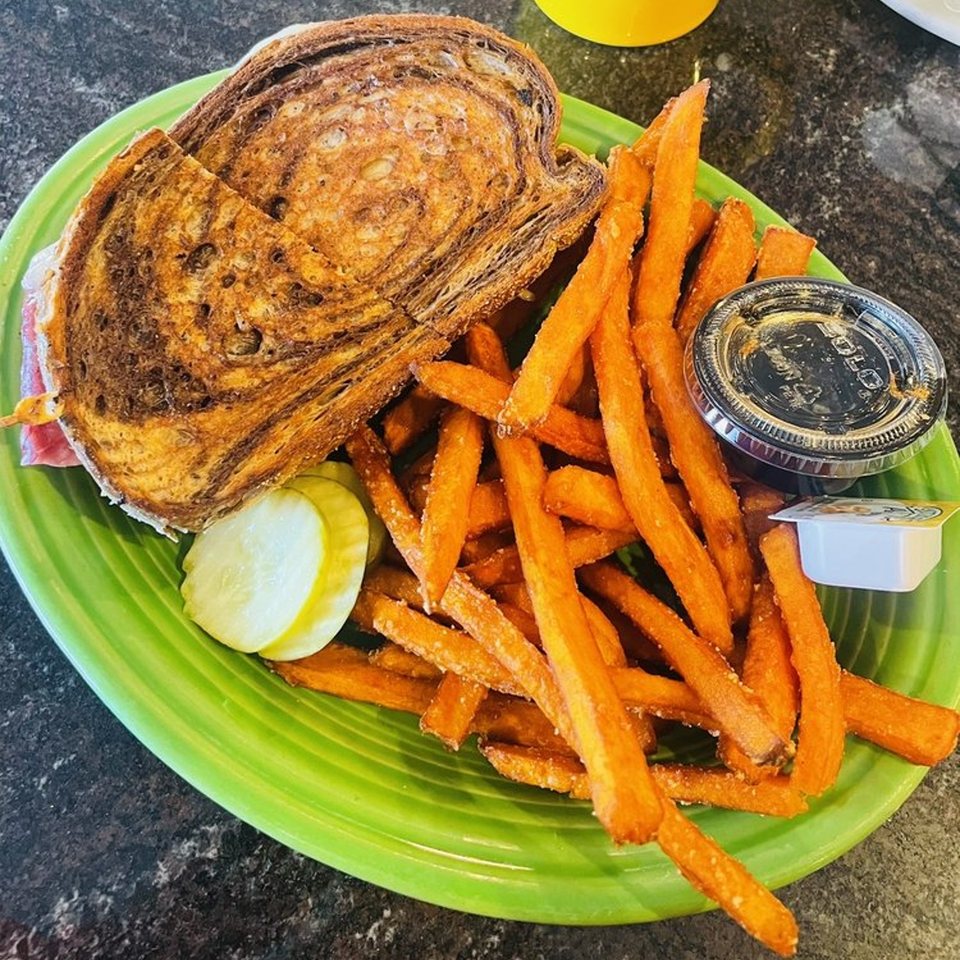 A sandwich on marbled rye bread with sweet potato fries and pickles on a green plate.